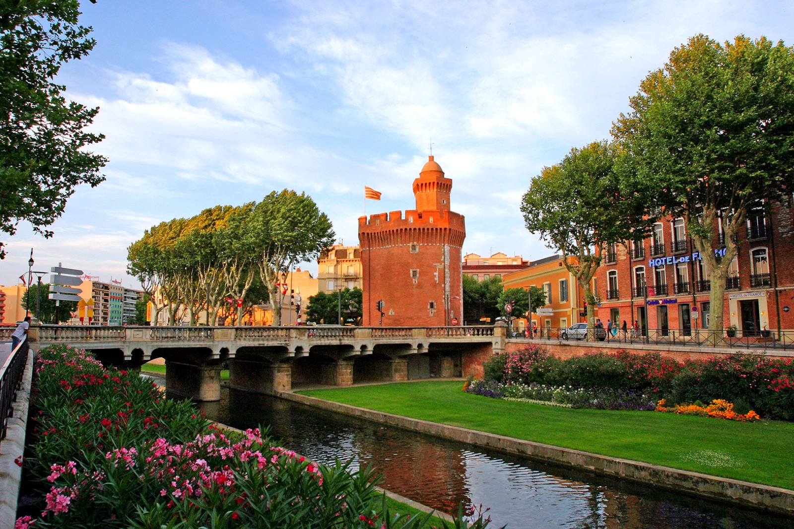 Le Castillet de Perpignan se refletant dans la Basse, avec les massifs de fleurs et les platanes du quai